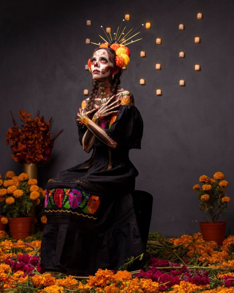 Woman in Catrina face paint and black dress surrounded by marigolds
